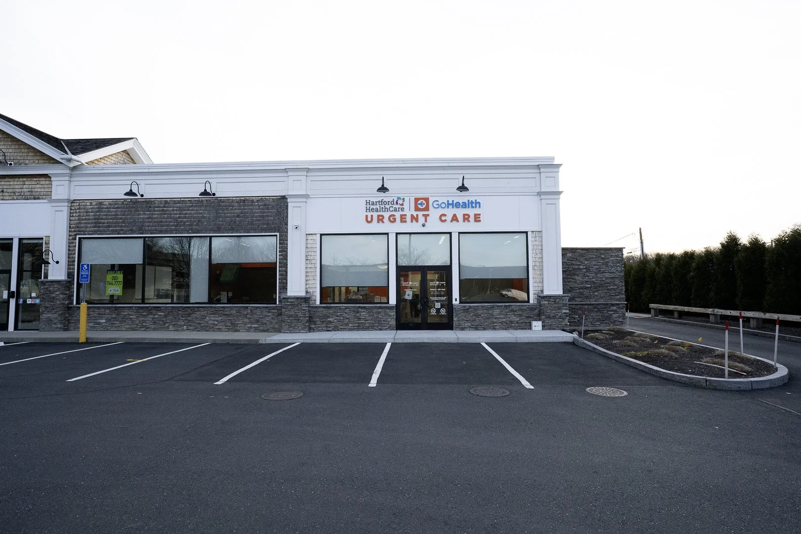 Front view of a modern medical urgent care clinic with parking spaces, signage for Hartford HealthCare GoHealth URGENT CARE, and a handicapped parking sign.