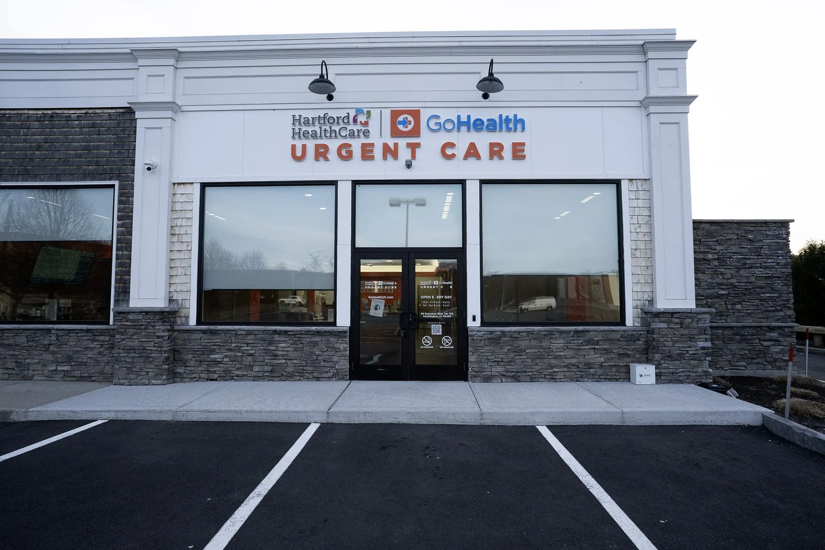 Front view of a healthcare urgent care clinic with signage for Hartford HealthCare GoHealth Urgent Care, large windows, and a parking lot in the foreground.