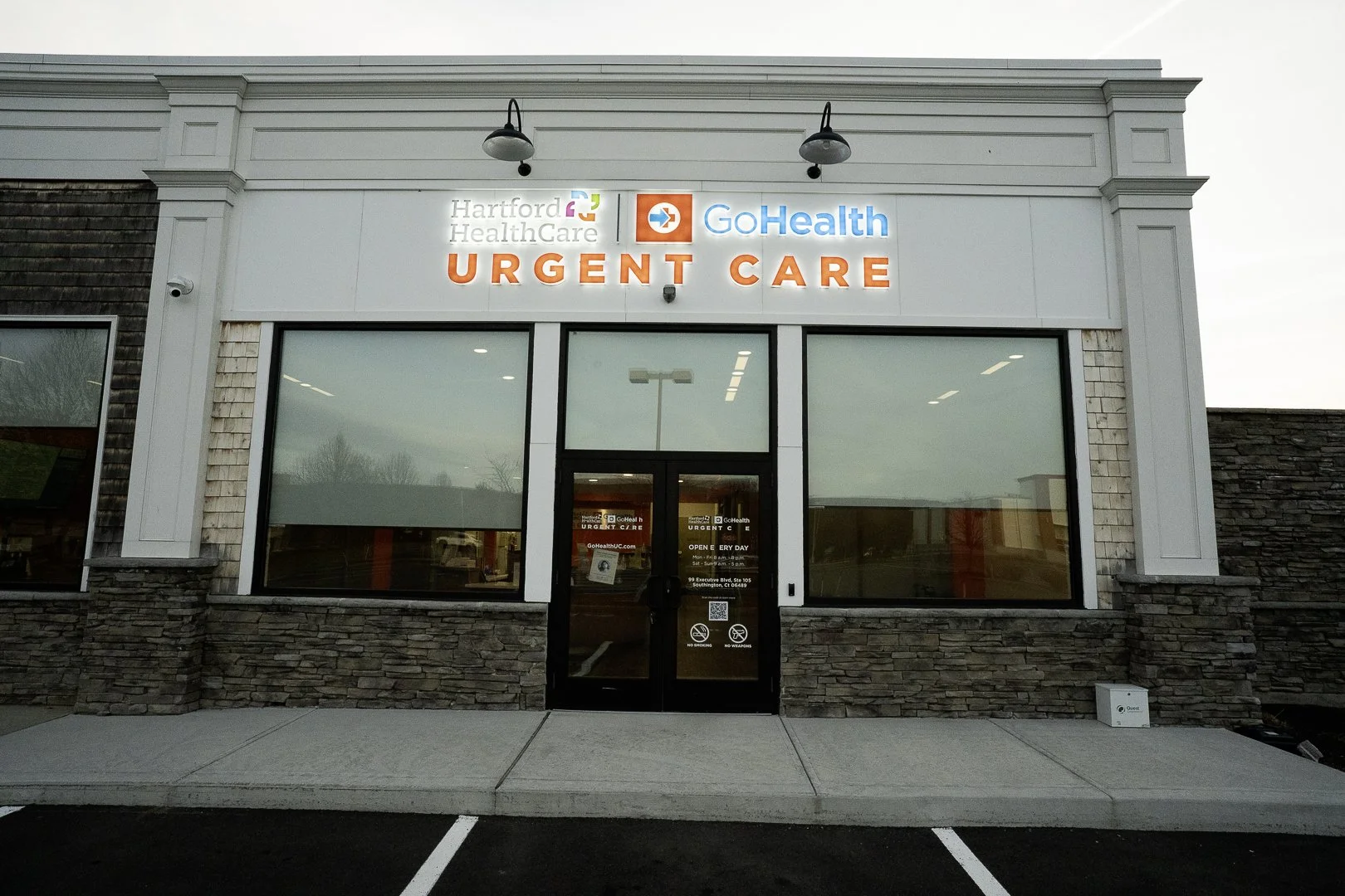 Exterior of a healthcare urgent care center with signs for Hartford Healthcare and GoHealth, featuring large windows, a double glass door, and parking spots in front.