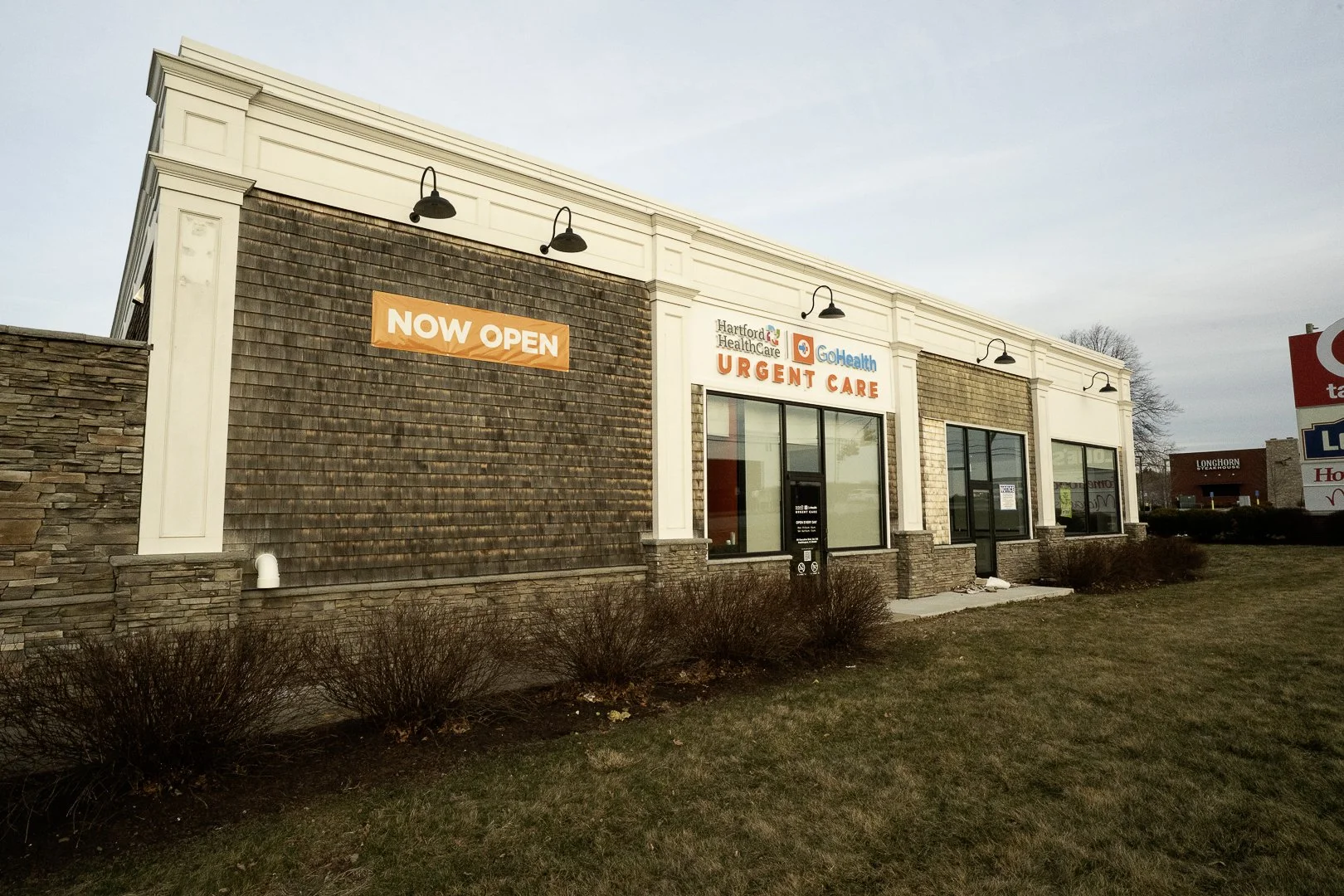 Exterior of a healthcare facility with signs indicating it is now open, offering urgent care services, with large windows and a brick facade.