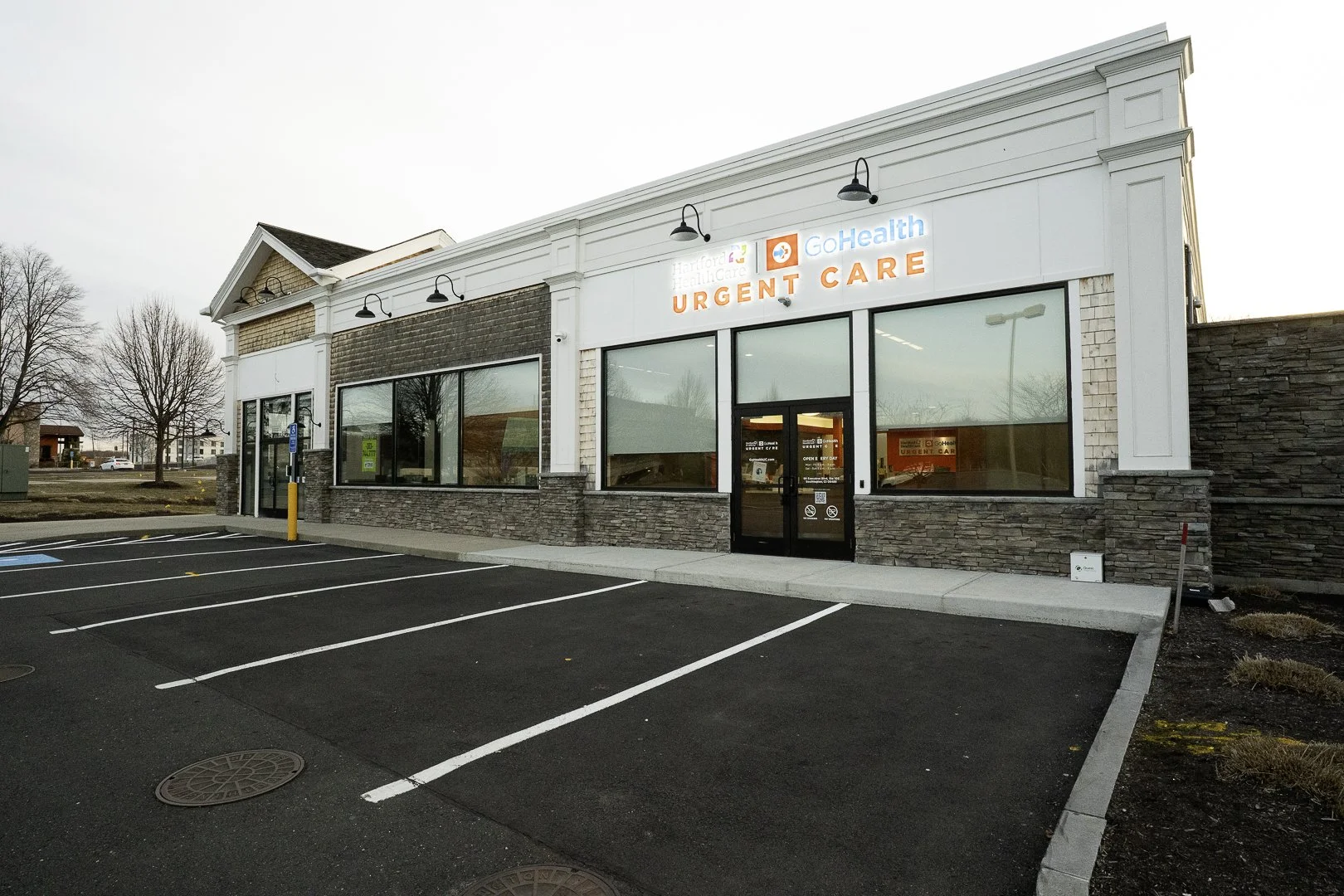 Exterior of a medical facility with signs for Hartford HealthCare, GoHealth, and Urgent Care, featuring large front windows, a black front door, and an empty parking lot.