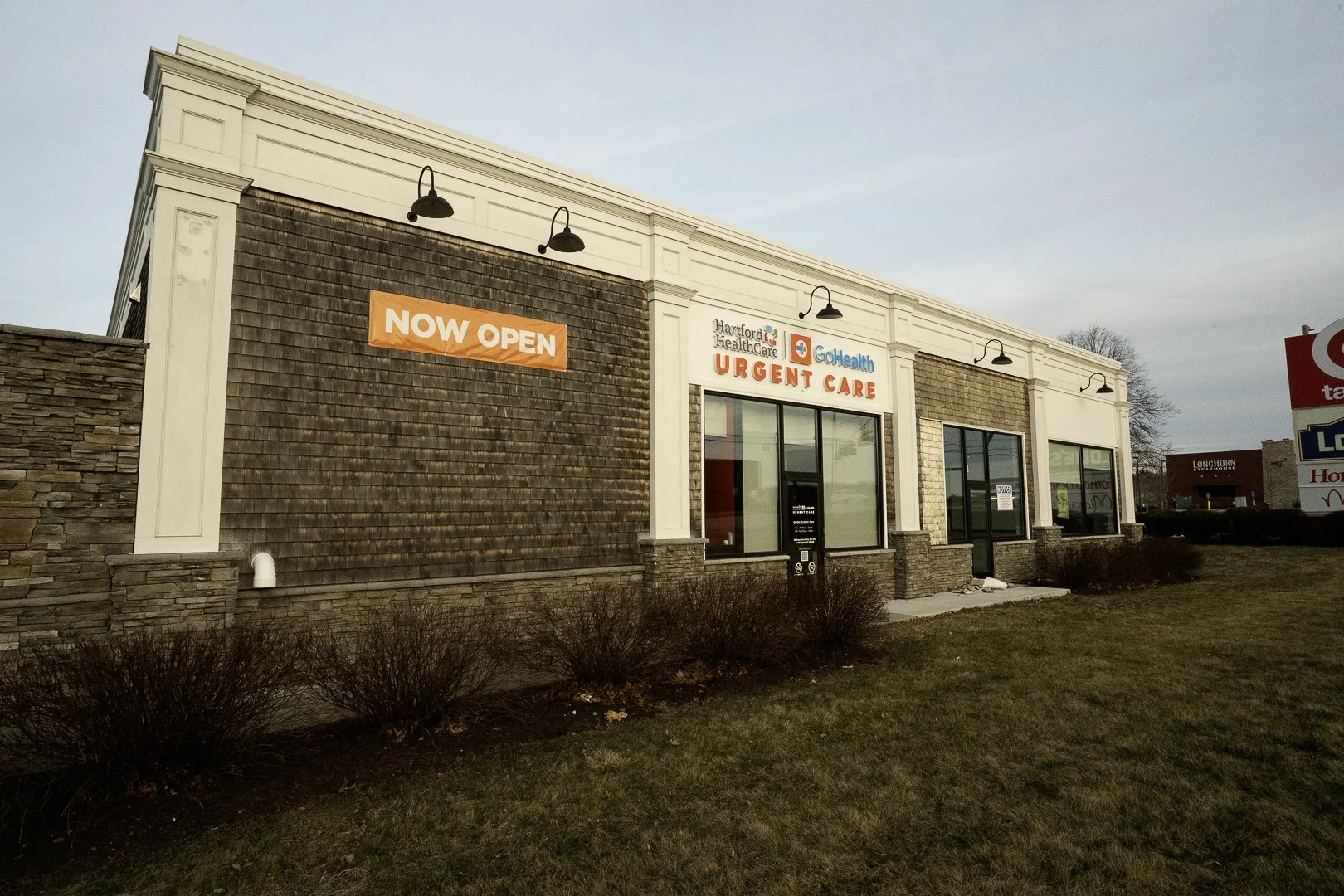 Exterior of an urgent care building with a sign that reads 'Now Open', under a partly cloudy sky. The building has a white trim, large windows, and a sign indicating it is part of Hartford Healthcare and GoHealth.