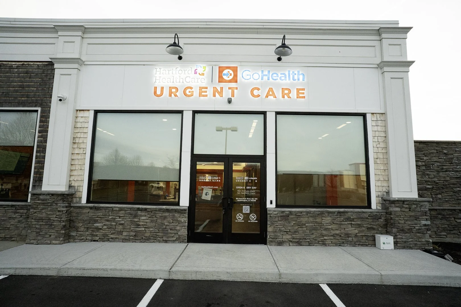 Exterior of an urgent care clinic with signs for Hartford HealthCare and GoHealth, featuring large windows, a glass door, and parking spaces in front.