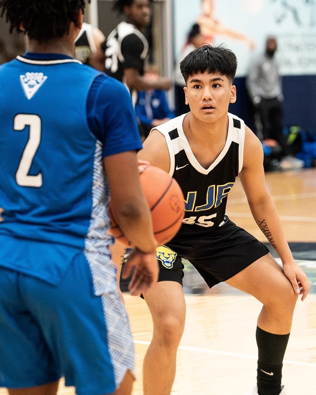 A basketball player in a black and white uniform with the number 45 and the logo NJF is defending against an opponent in a blue uniform with the number 2, holding a basketball during a game.