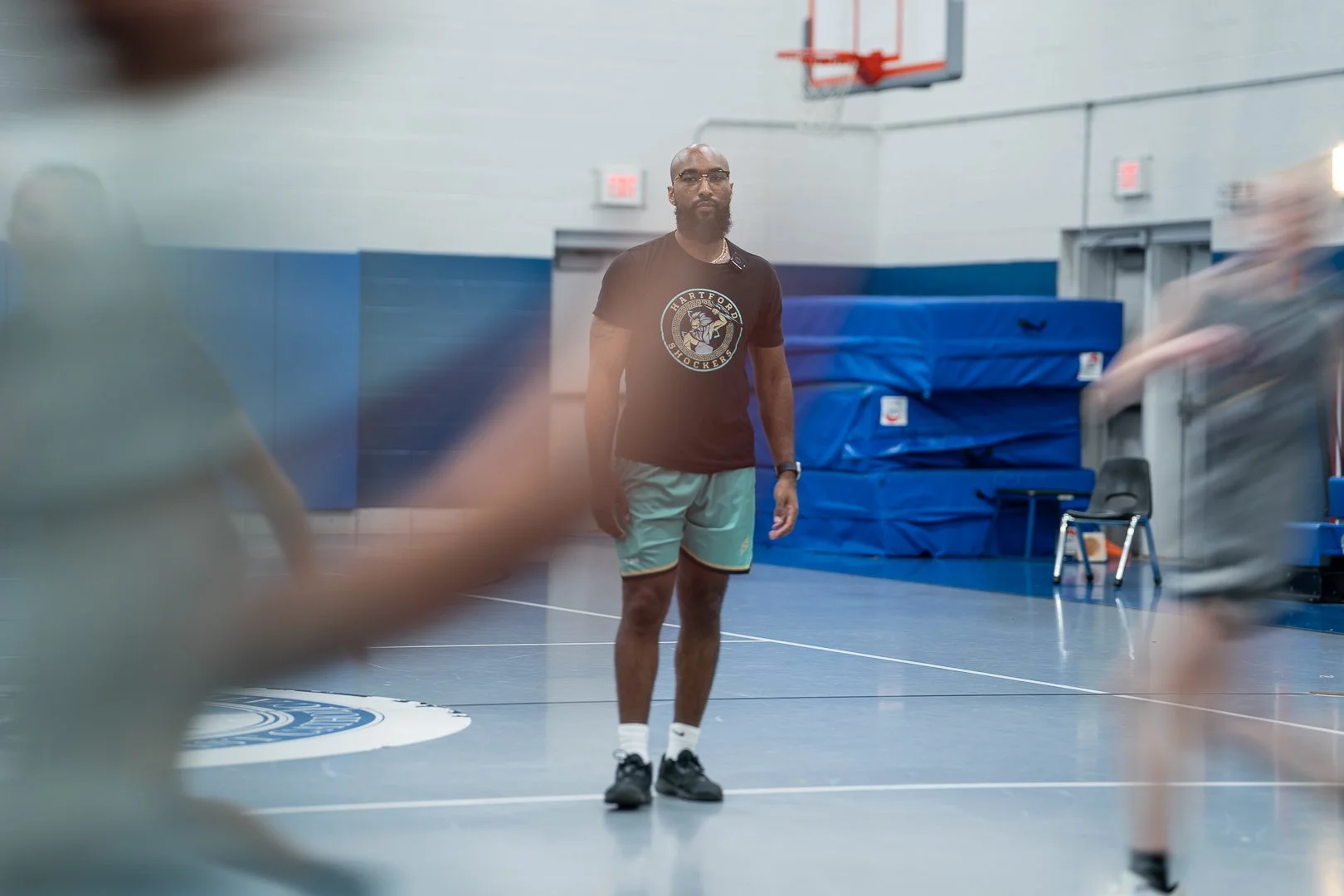 Man standing in a gymnasium, wearing a black T-shirt with a Hartford Hockey logo, green shorts, and black sneakers, surrounded by blurred figures.