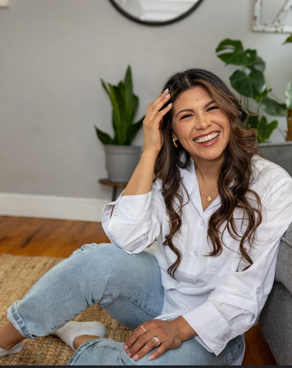 A woman sitting on the floor, smiling and touching her hair. She is wearing a white shirt and blue jeans, with plants and a clock in the background.