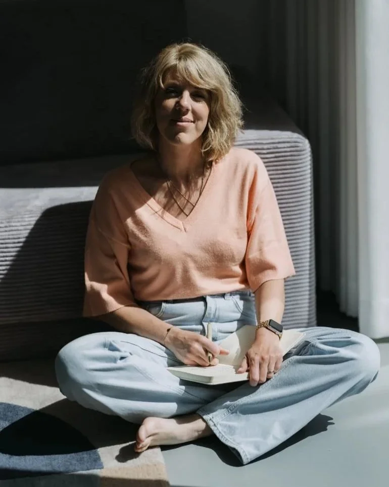 Vrouw zittend op de vloer met een boek, binnen, met zonlicht dat door de ramen schijnt.