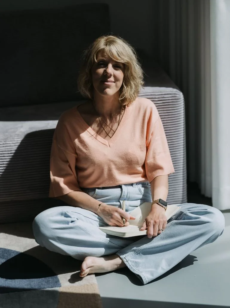 Vrouw zit op de vloer met gekruiste benen, schrijft in een boek, draagt een roze overhemd en witte spijkerbroek, en kijkt naar de camera met een glimlach, met zonlicht dat door een raam op haar schijnt.
