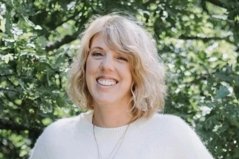 Een lachende vrouw met blond, krullend haar poseert voor groene bomen in de achtergrond.