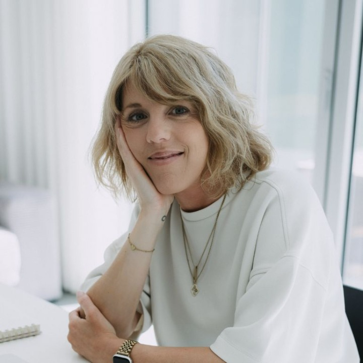 Een vrouw met schouderlang, golvend blond haar, zittend aan een bureau, rust op haar wang met haar hand, glimlachend.
