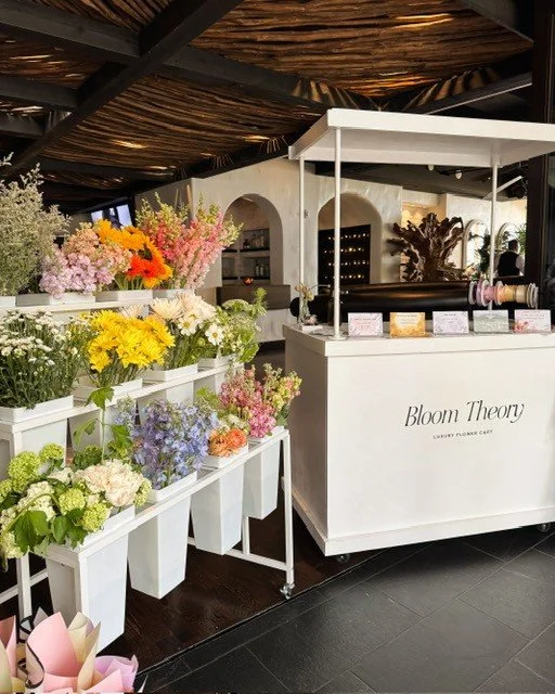 A flower stand with colorful fresh flowers and a white cart labeled 'Bloom Theory' at an indoor market.
