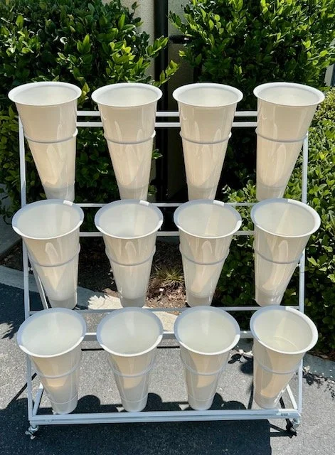 White buckets arranged on a three-tier metal flower display rack outdoors with green bushes in the background.