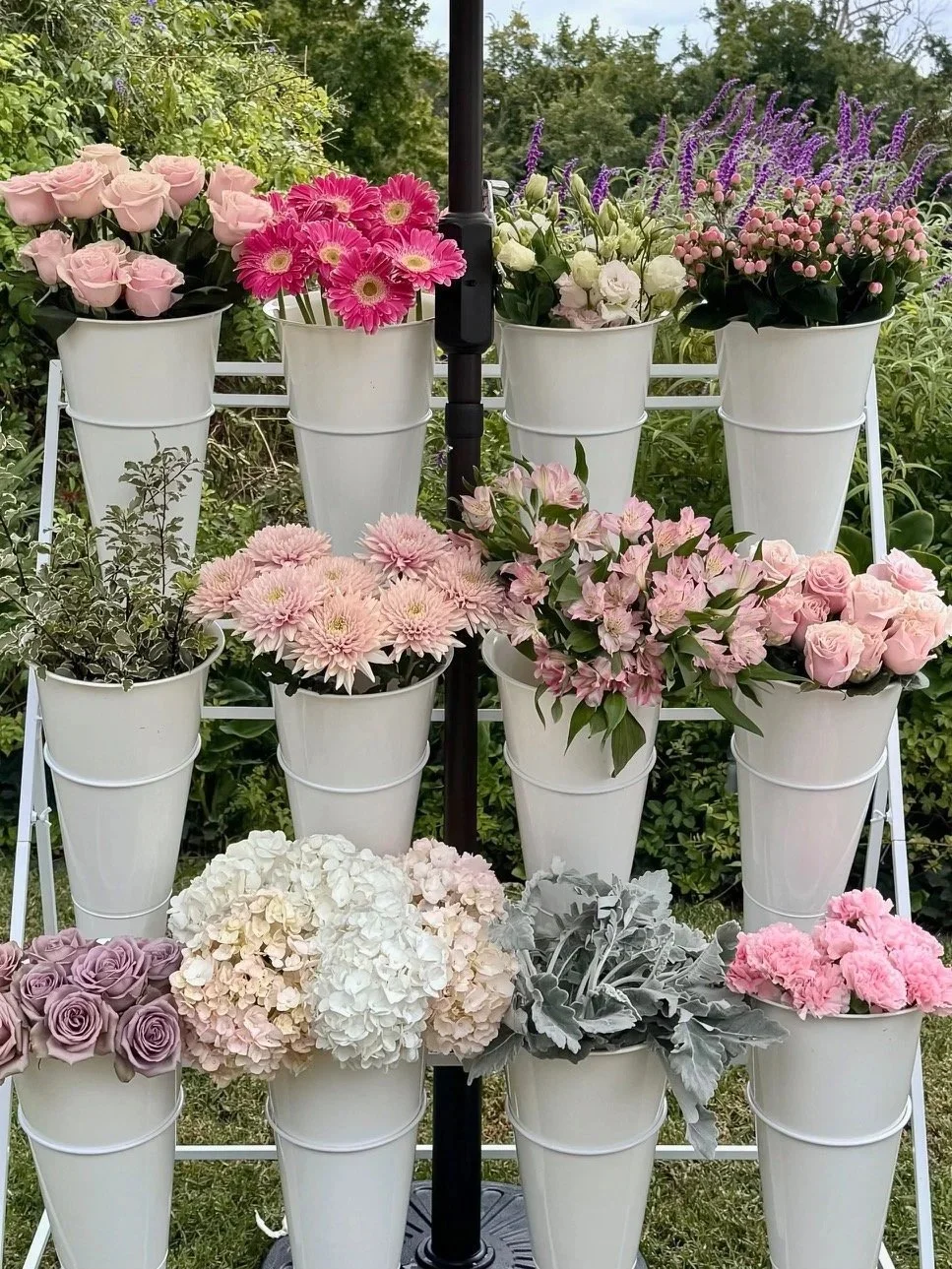 Display of various pink, purple, and white flowers in white buckets on a metal flower bouquet stand outdoors.