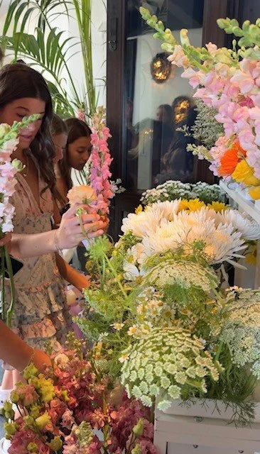 Two girls shopping for flowers at a floral shop with various colorful flowers and greenery on display.