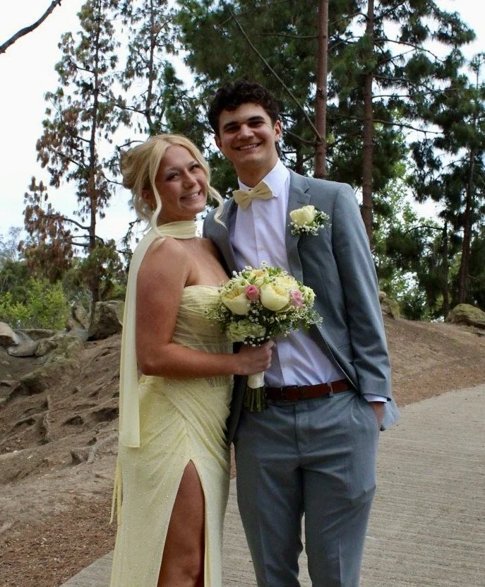 A bride and groom standing outdoors in a wooded area, smiling at the camera. The bride is wearing a light yellow strapless gown with a thigh-high slit, and is holding a bouquet of white and pink flowers. The groom is dressed in a gray suit with a white shirt, yellow bow tie, and a white boutonnière.