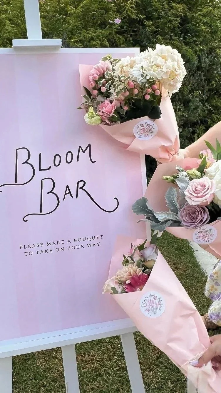 A pink Bloom Bar sign on an easel reads "Bloom Bar" with the message "Please make a bouquet to take on your way." Several bouquets of assorted flowers wrapped in pink paper are displayed next to the sign, set outdoors with a green shrub background.