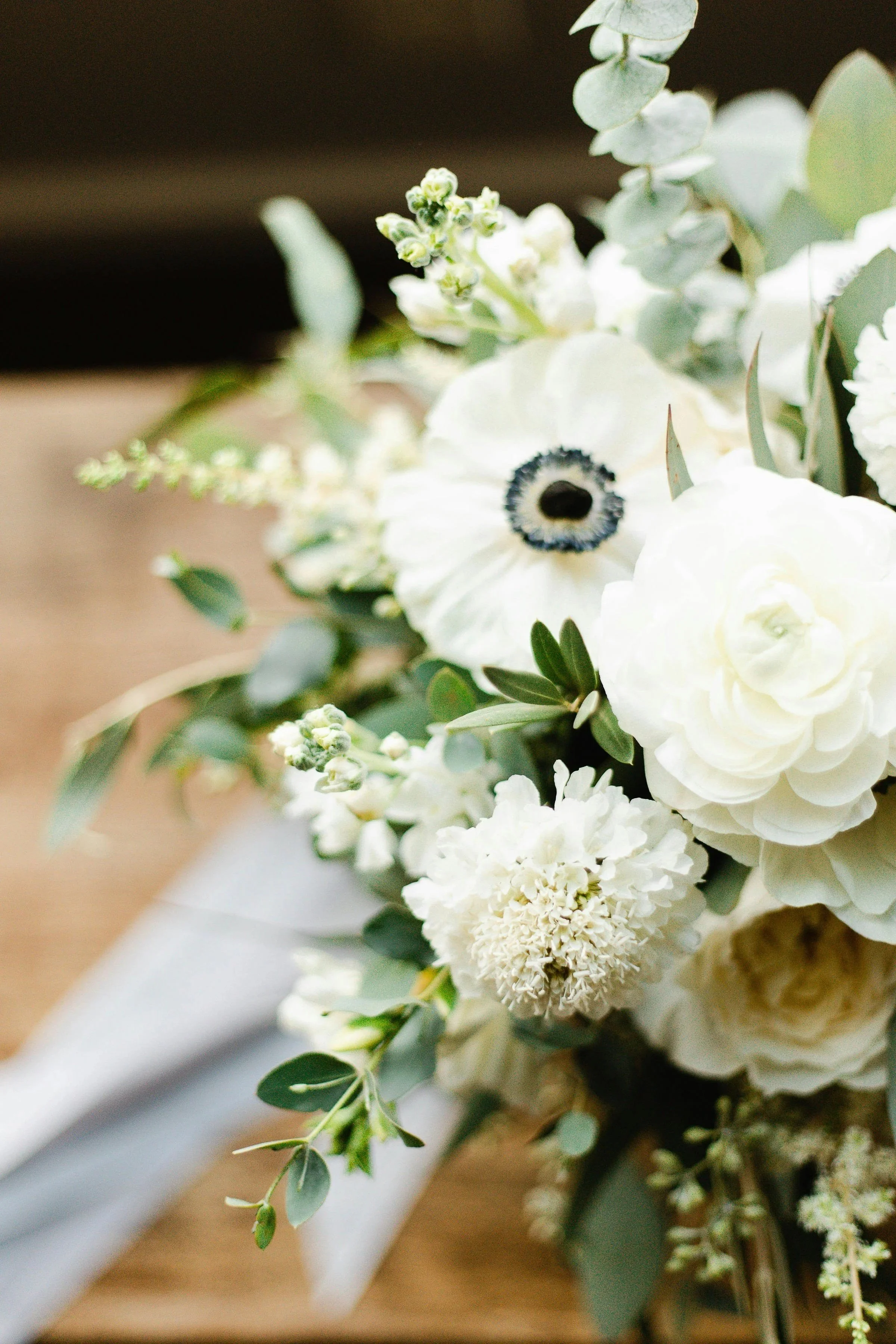 tied flowers, anemone, ranunculus, peonies. Close-up of a white bouquet with various flowers and green foliage, featuring a large white flower with a dark center, set against a soft background.