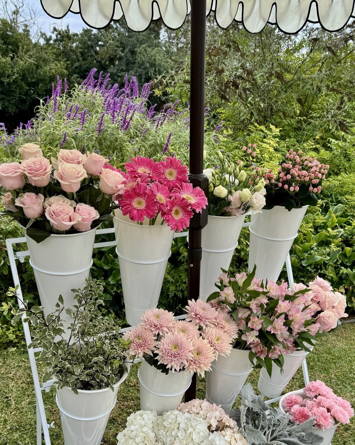 Display of various pink, purple, and white flowers in white buckets arranged on a metal stand outdoors with greenery and trees in the background. Bloom bar setup for baby shower in Newport Beach