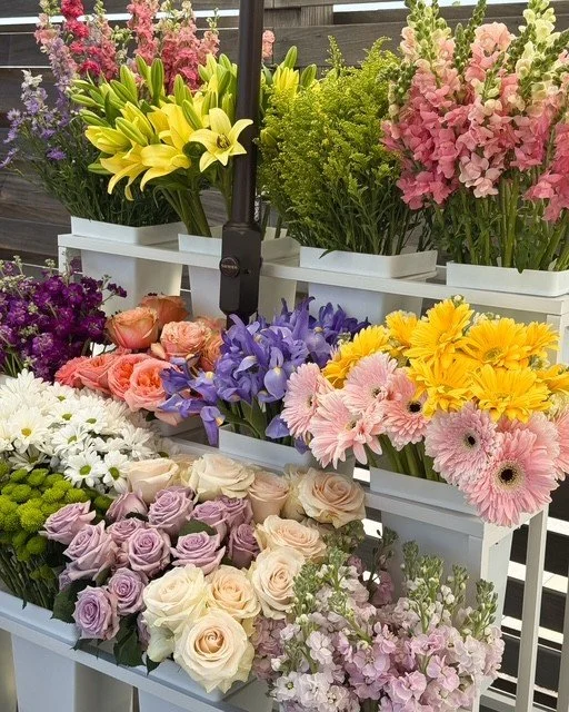 Bloom Bar, flower bar, orange county, A bouquet of pink, white, and purple flowers wrapped in pink paper, placed on a white flower cart outside with green and purple plants in the background.