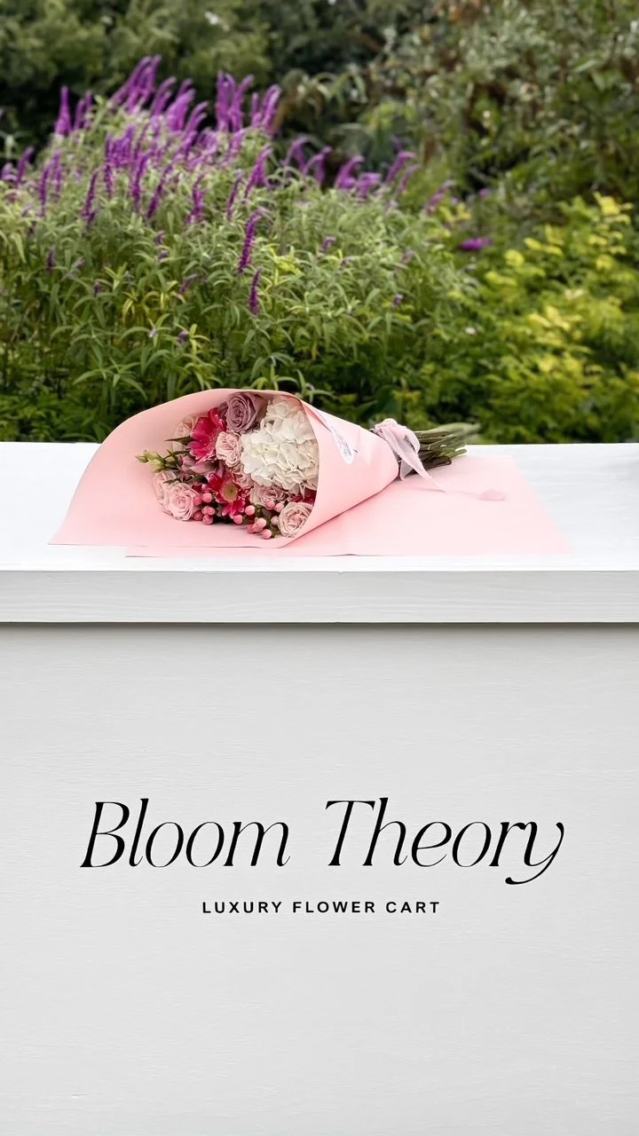 A bouquet of pink, white, and purple flowers wrapped in pink paper, placed on a white flower cart outside with green and purple plants in the background.