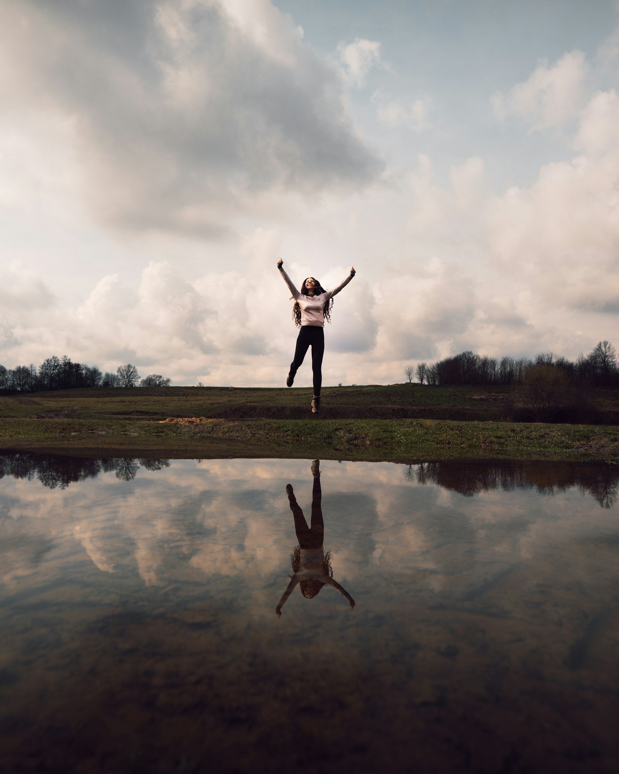 A woman jumping with arms raised on a grassy field, with a cloudy sky and trees in the background, and her reflection in a body of water below.