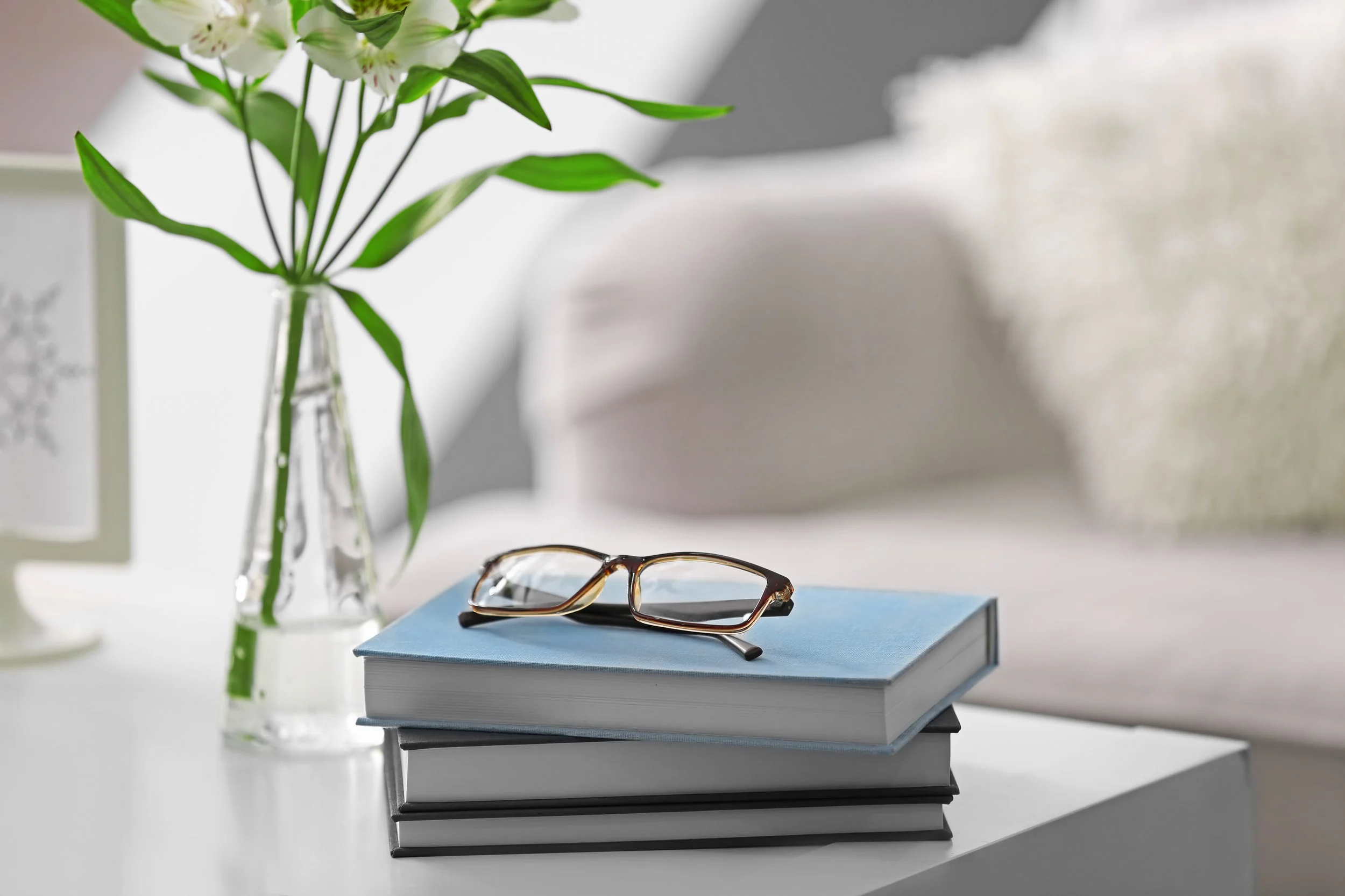 A stack of books with a pair of glasses on top, a vase with green leaves and white flowers, and a blurred background with neutral tones
