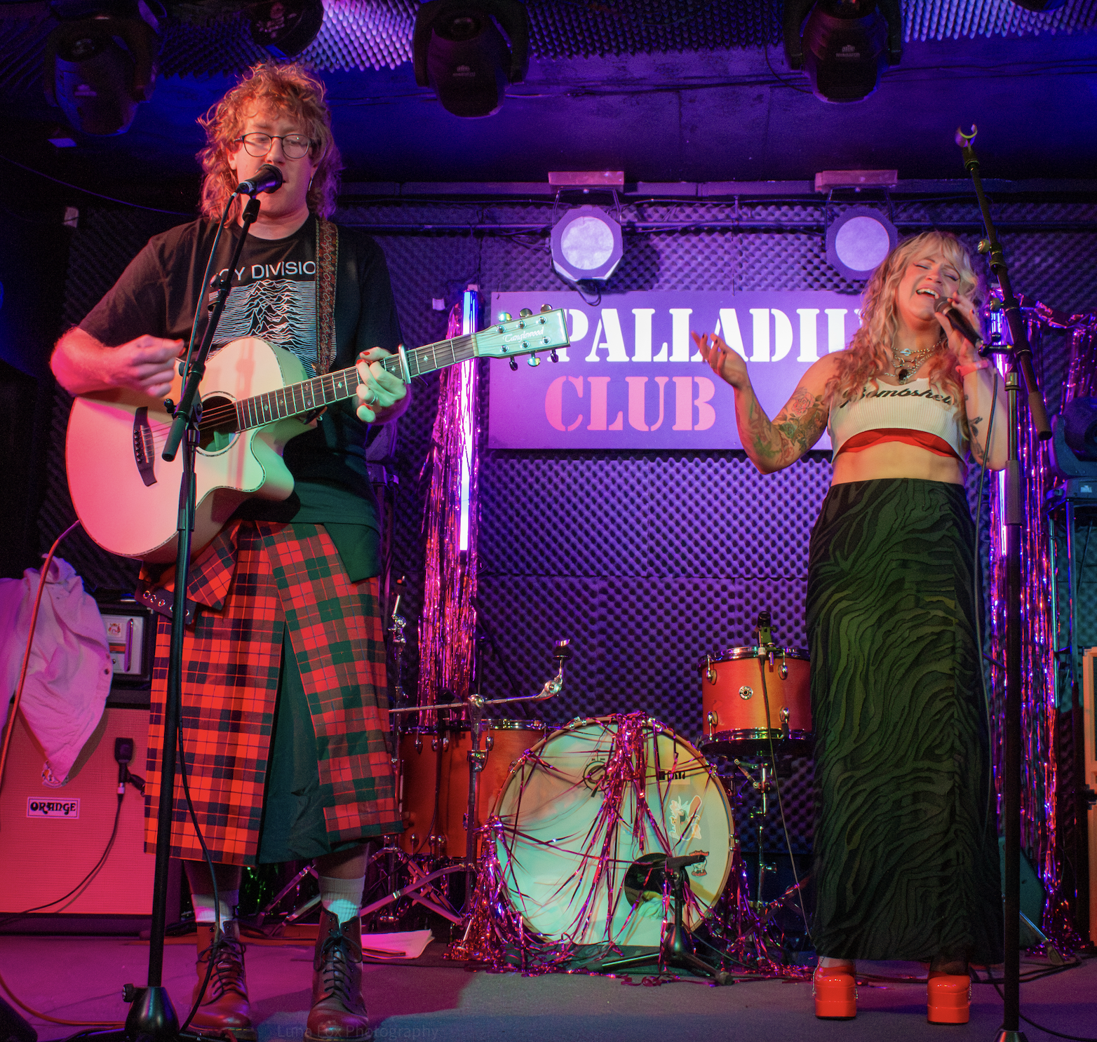 A musician playing an acoustic guitar on stage with a singer, performing at the Palladium Club. The stage is decorated with purple and pink lights, and there is a drum set behind them. The musician is wearing glasses, a black t-shirt, a red plaid ski