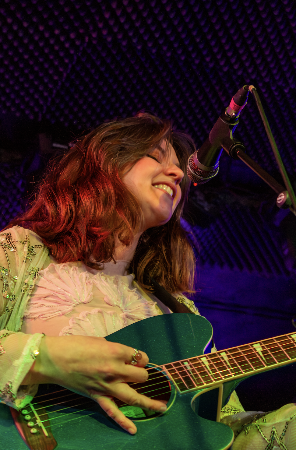 A woman with wavy brown hair, smiling and singing into a microphone while playing an acoustic guitar with a dark green finish, in a recording studio with soundproof foam ceiling.