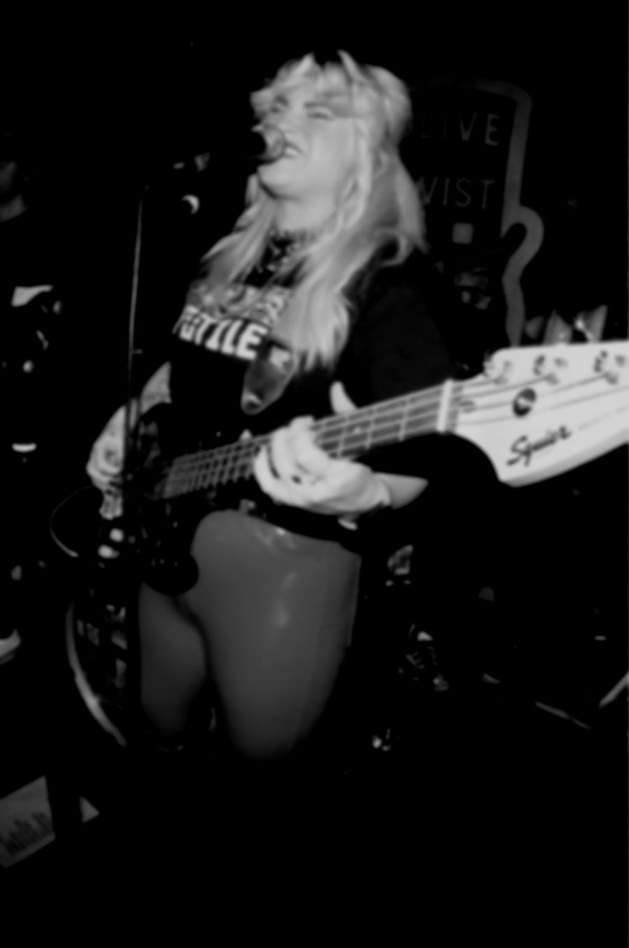 A woman singing and playing an electric bass guitar in a dimly lit music venue.