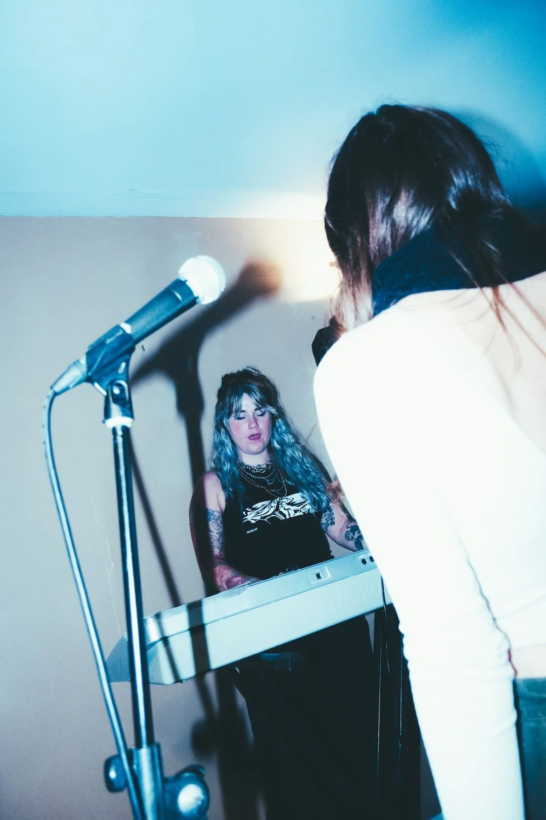 Person with blue hair and tattoos performing on a keyboard, with a microphone nearby, in a room with shadowed plain wall. Singing Lessons Bideford, Music Teacher Bideford, Singing Lessons North Devon, Bold Note Vocal Coaching.