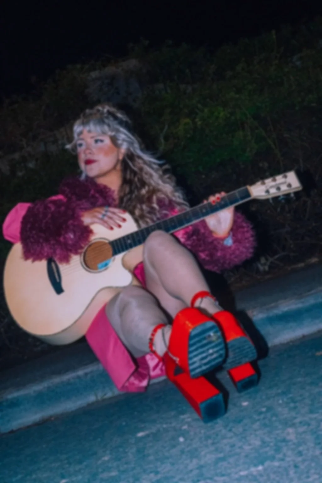 A woman with long curly hair and makeup, wearing a fuzzy maroon sweater, is sitting on the sidewalk at night playing an acoustic guitar. She has bright red shoes and is surrounded by dark foliage in the background. Bold Note Vocal Coaching