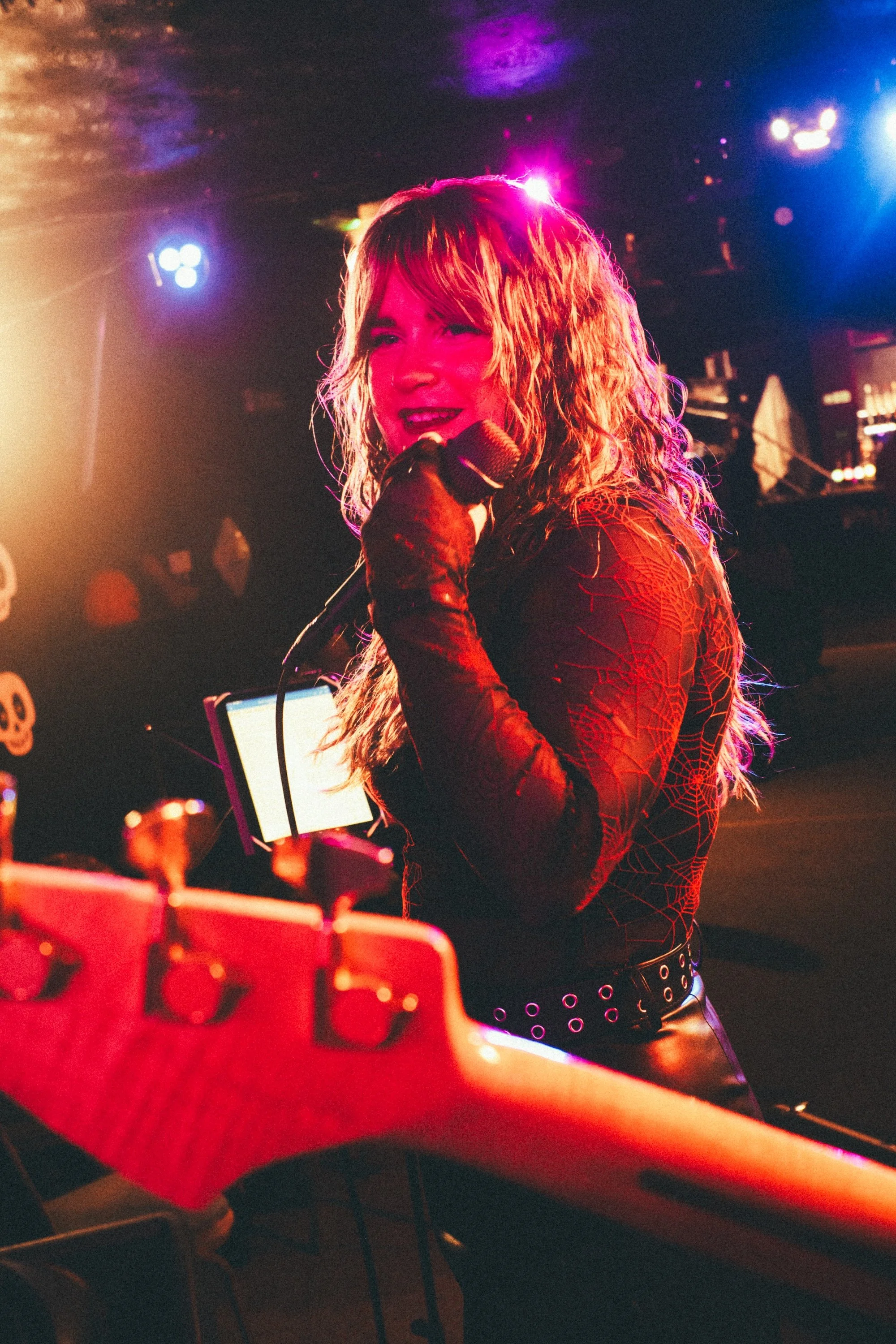A woman with wavy hair singing into a microphone on stage, illuminated by colorful stage lights, with musical equipment and a guitar in the foreground. Singing Lessons Bideford, Music Teacher Bideford, Singing Lessons North Devon, Bold Note Vocal