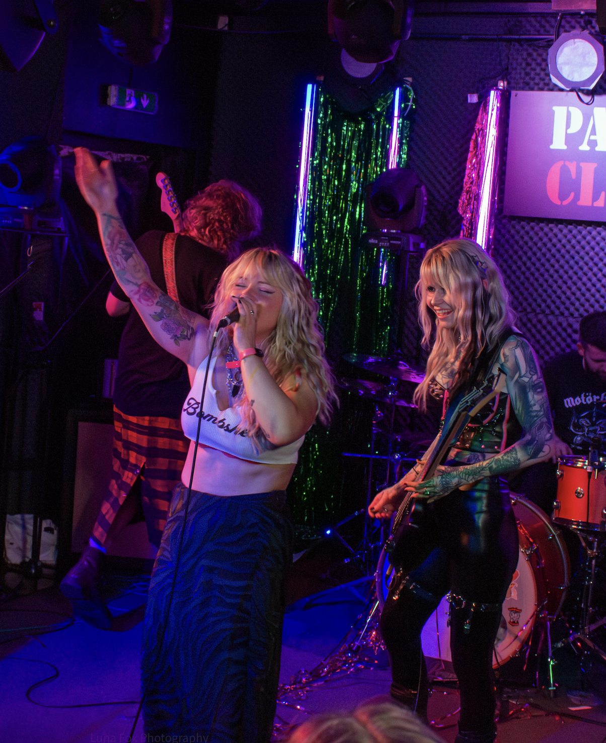 Two women with tattoos performing on a colorful stage with neon lights, one singing into a microphone and the other playing guitar, with a drummer in the background.