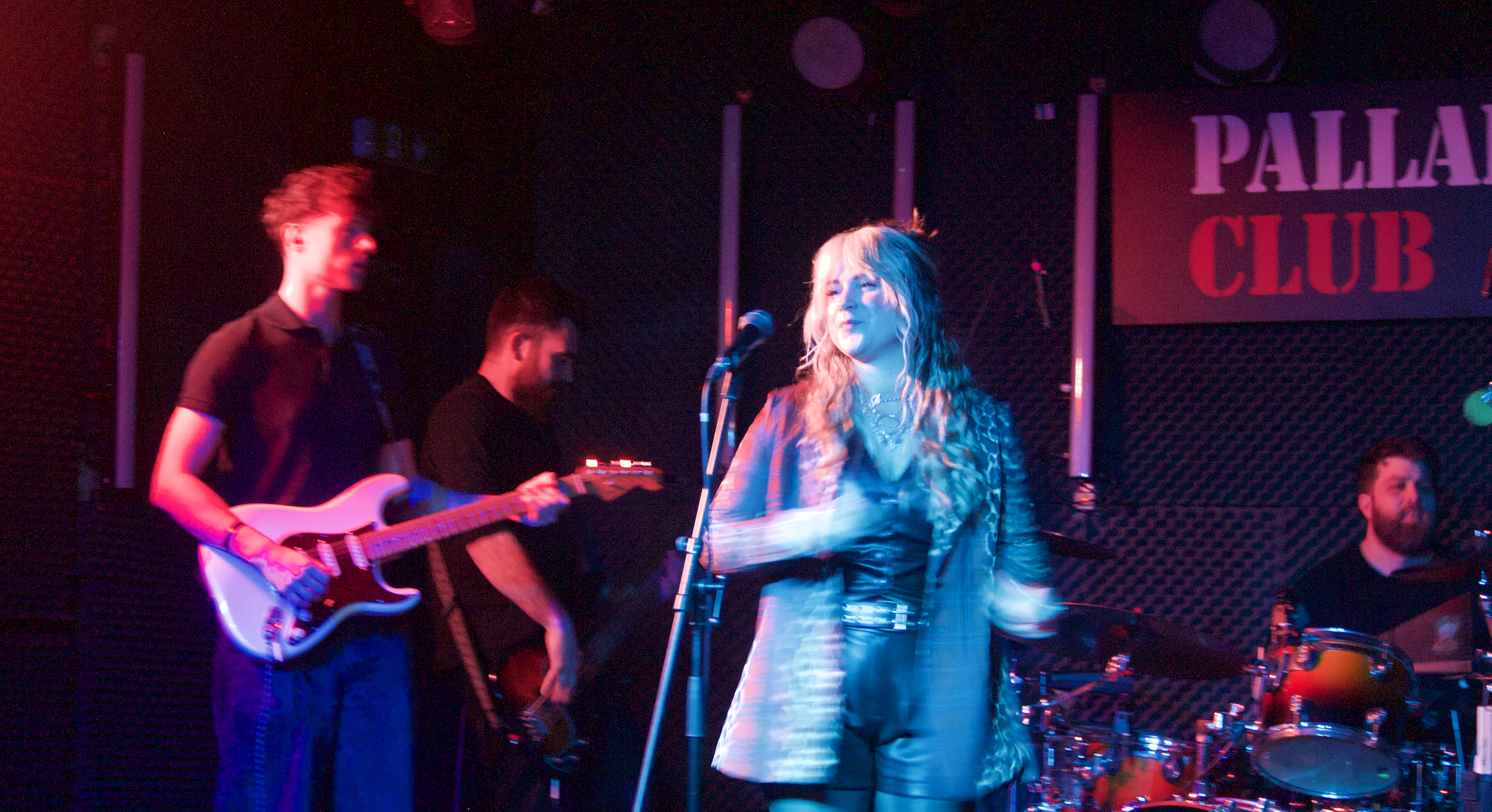 A band performing on stage at Palladium Club, with a female singer in the center, a guitarist to her left, another musician in the background, and a drummer on the right, all under colorful stage lighting.