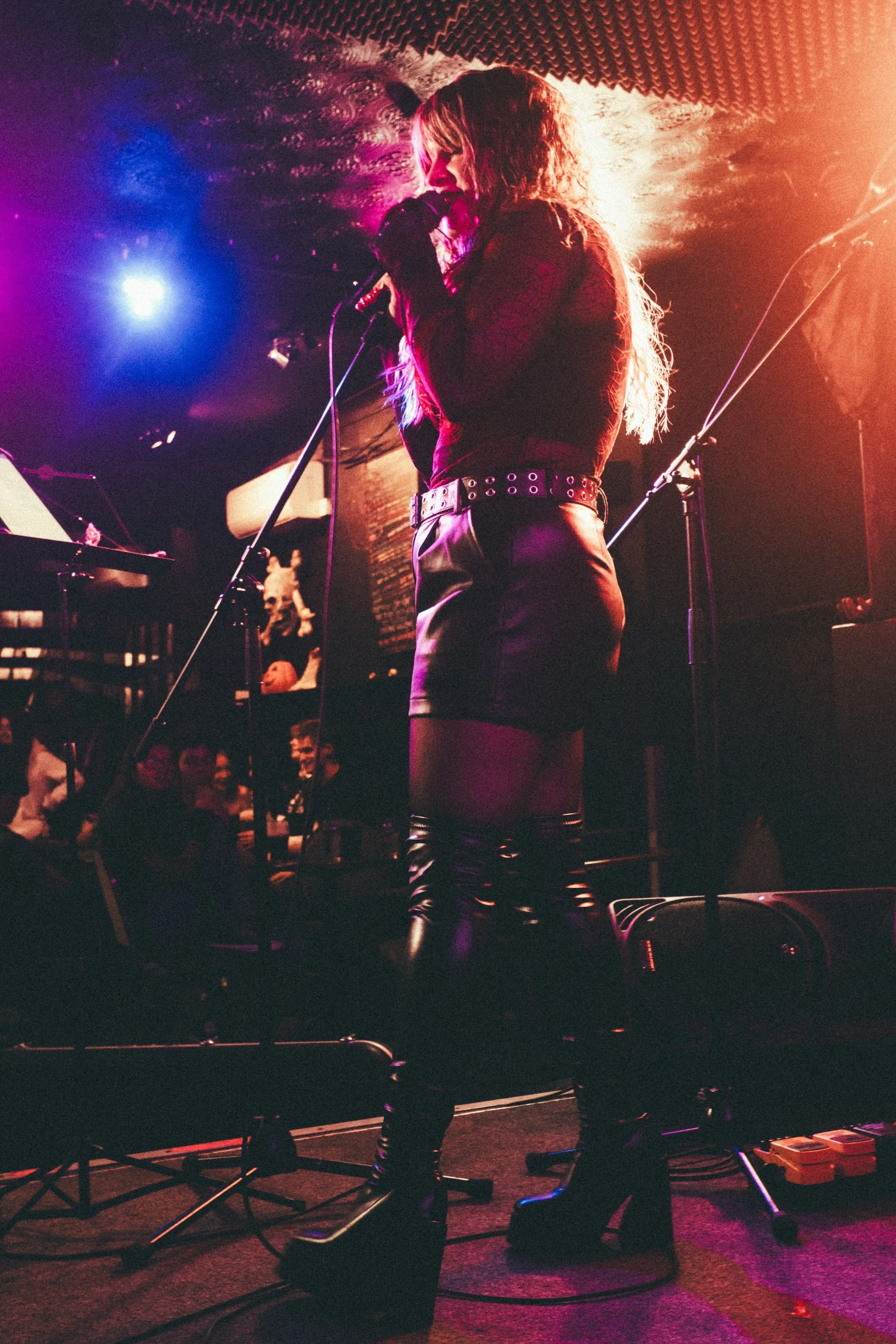 A female singer with long curly hair performing on stage in a dimly lit venue, wearing black leather shorts, and a lace top, with an audience visible in the background. Singing Lessons Bideford, Music Teacher Bideford, Singing Lessons North Devon, Bo
