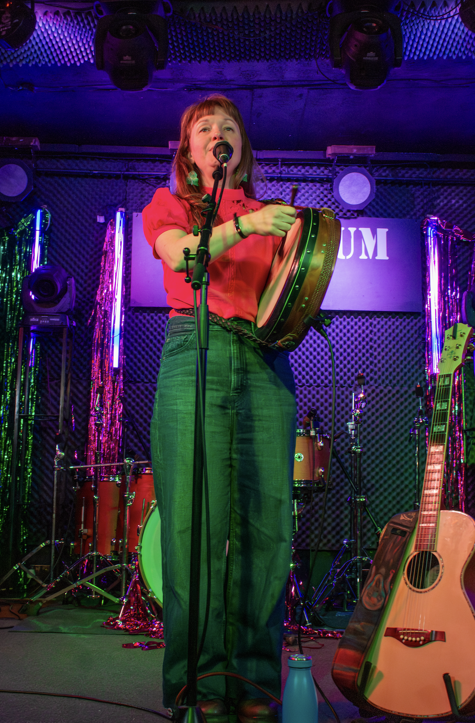 A woman singing into a microphone while holding a tambourine on a stage lit with colorful neon lights, with musical instruments including an acoustic guitar and drums in the background.