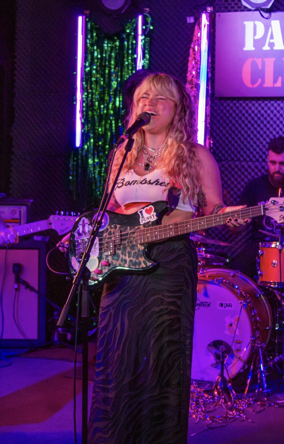 A woman singing into a microphone and playing an electric guitar on stage with colorful neon lights and a drum set in the background.