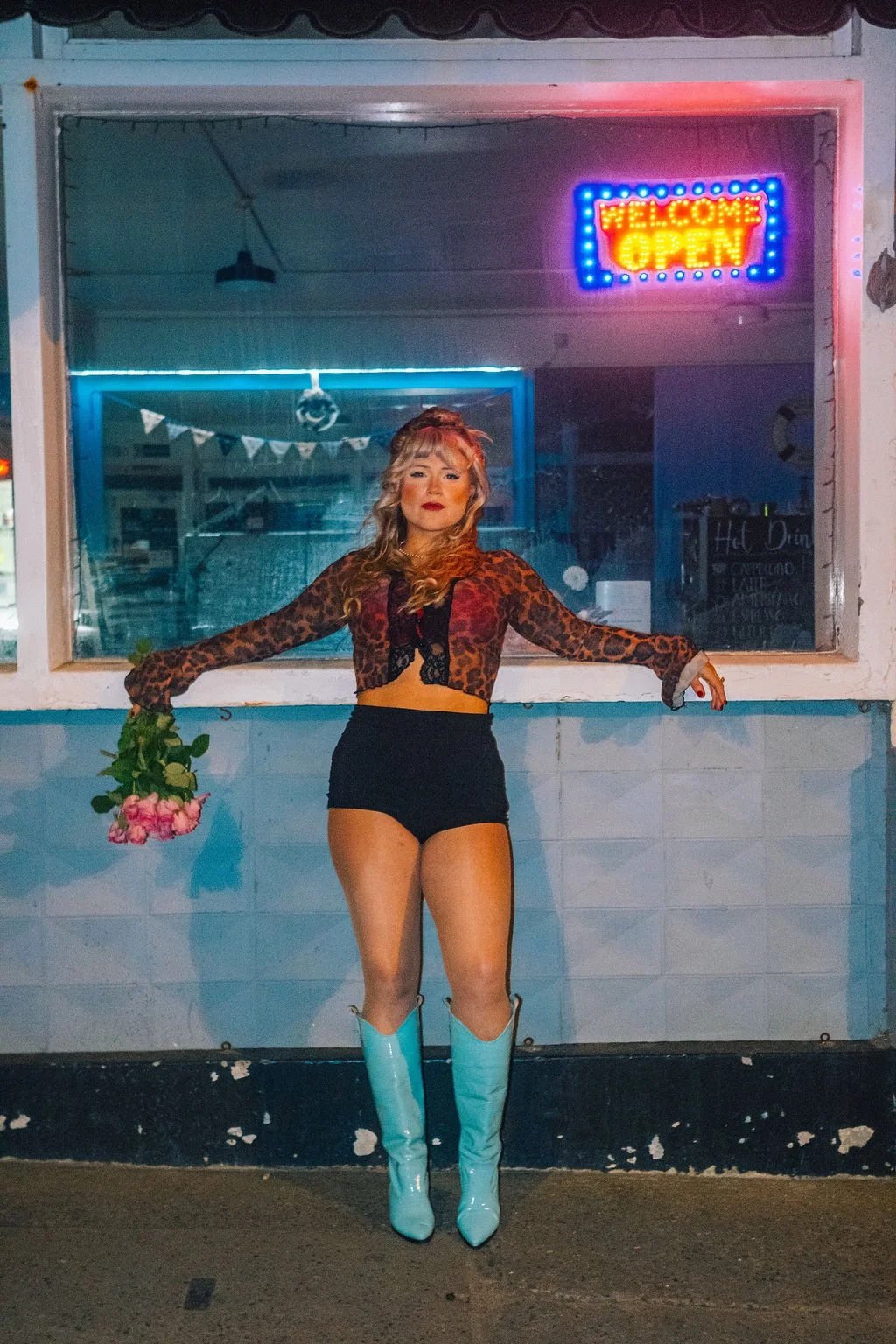 Woman standing in front of a window with neon signs, holding pink flowers, wearing a leopard print top, high-waisted shorts, and light blue cowboy boots, at night.