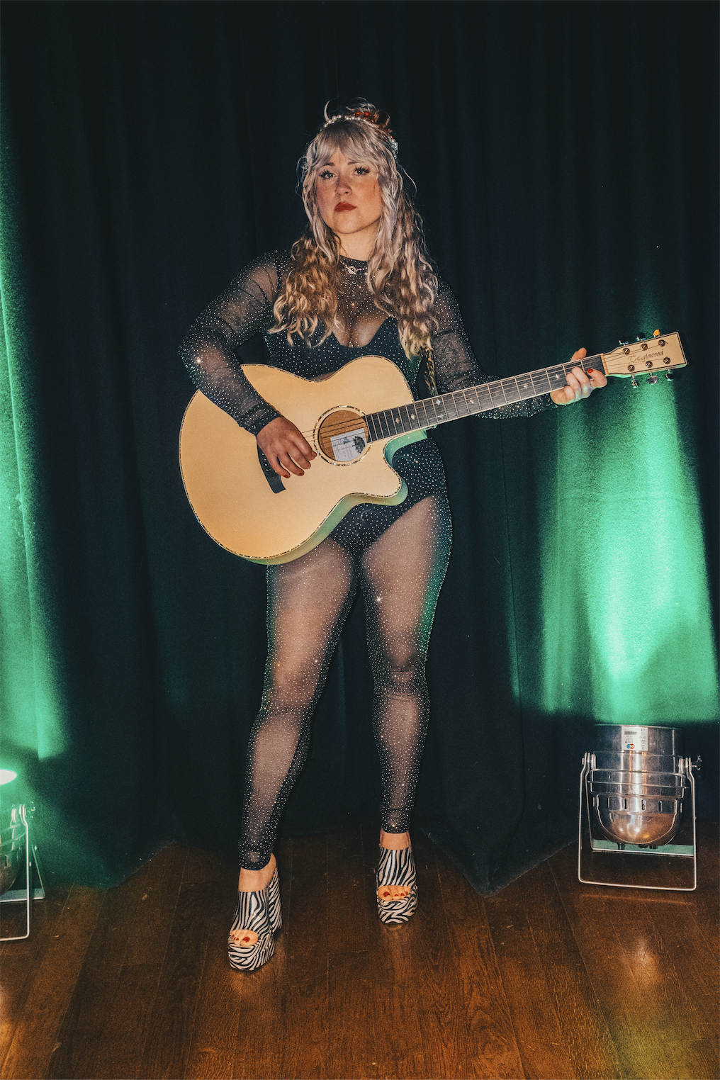 A woman with long wavy hair, wearing a sheer black outfit with sparkles and zebra-print high heels, playing an acoustic guitar on a stage with black curtain and green lighting.