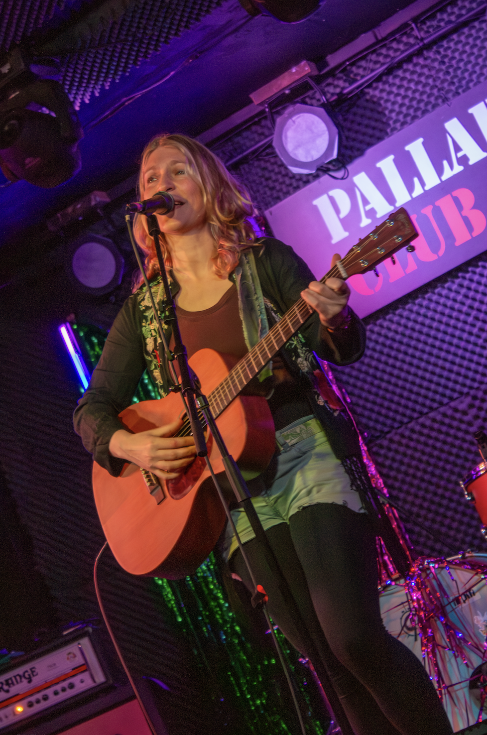 A woman with wavy, shoulder-length hair plays an acoustic guitar and sings into a microphone on stage at the Palace Club, with stage lights and a sign in the background.