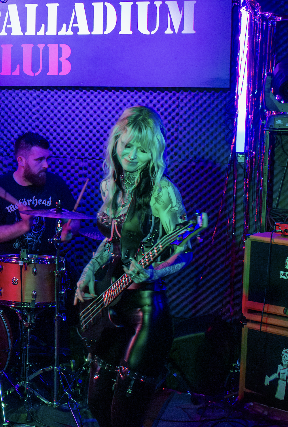 A woman playing a bass guitar and smiling on stage with a drummer in the background at alladium club, illuminated by colorful stage lights.