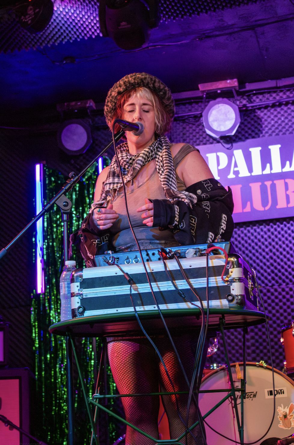 A female musician with blonde hair, wearing a leopard-print hat, fishnet stockings, and layered clothing, performs on stage with a microphone and electronic equipment. The background features colorful stage lighting and a sign that reads 'PALLA CLUB'