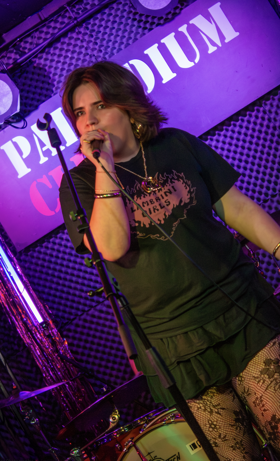 A young woman singing into a microphone on stage at a live music venue, with purple and pink lighting, wearing a black T-shirt with a graphic design, a necklace, bracelets, a black skirt, and fishnet stockings.
