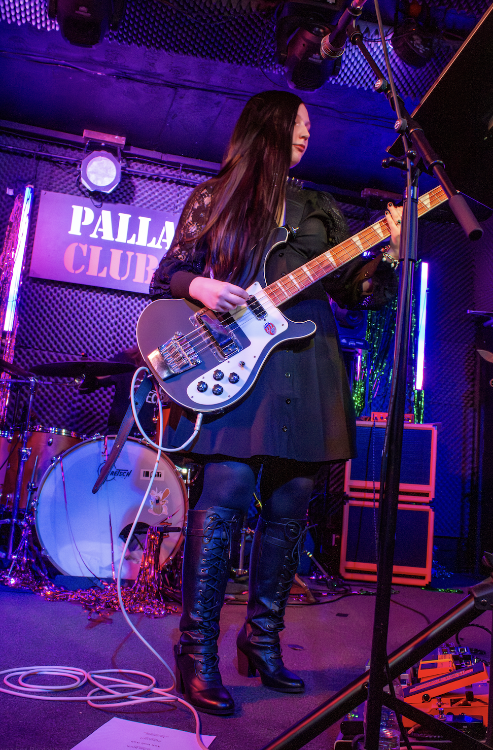 A woman playing bass guitar on stage at Pallas Club, with drums and colorful lighting in the background.