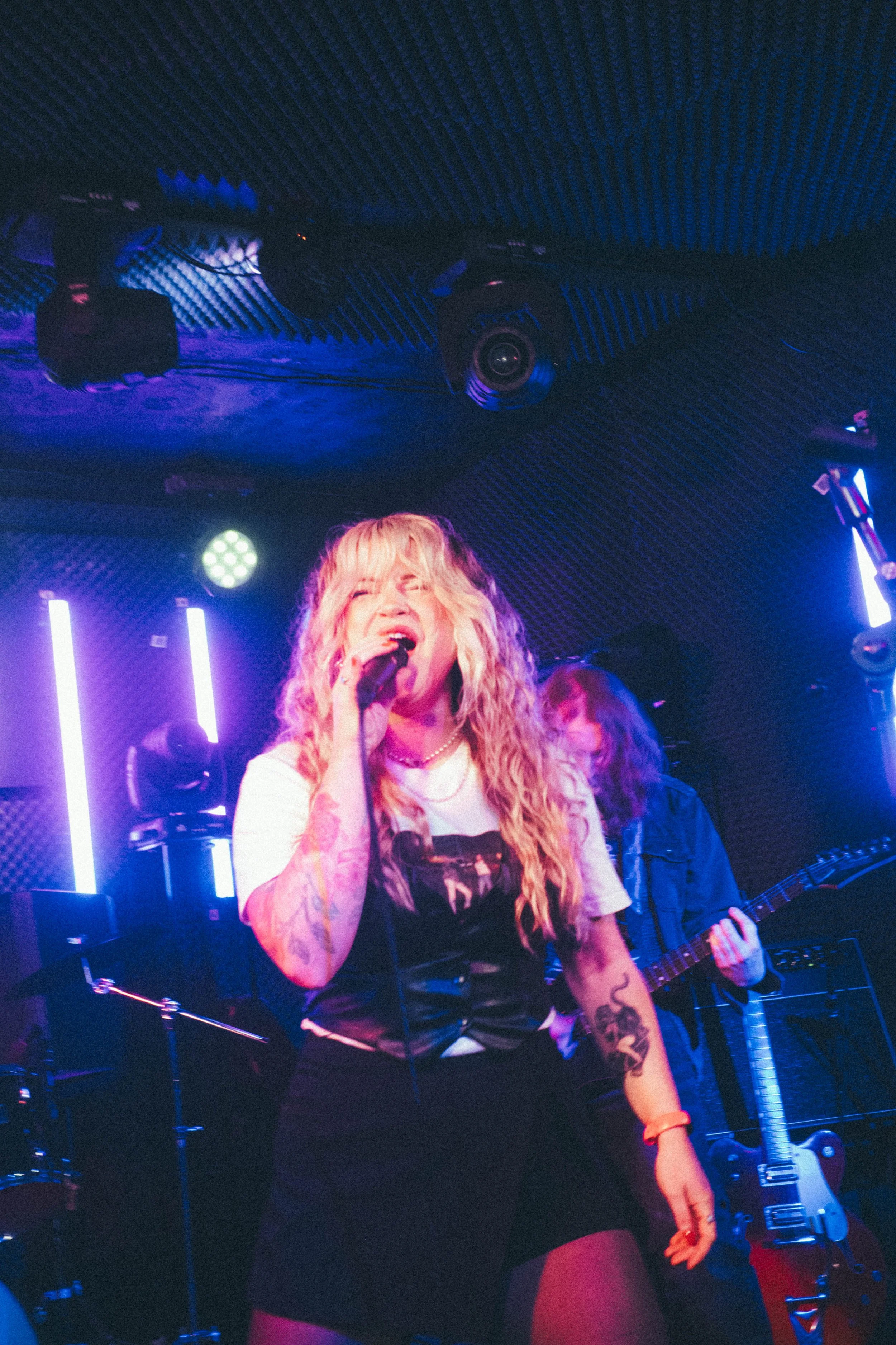 Female singer with long, curly blonde hair singing into a microphone on stage, accompanied by a guitarist, with colorful stage lights and sound equipment in the background.
