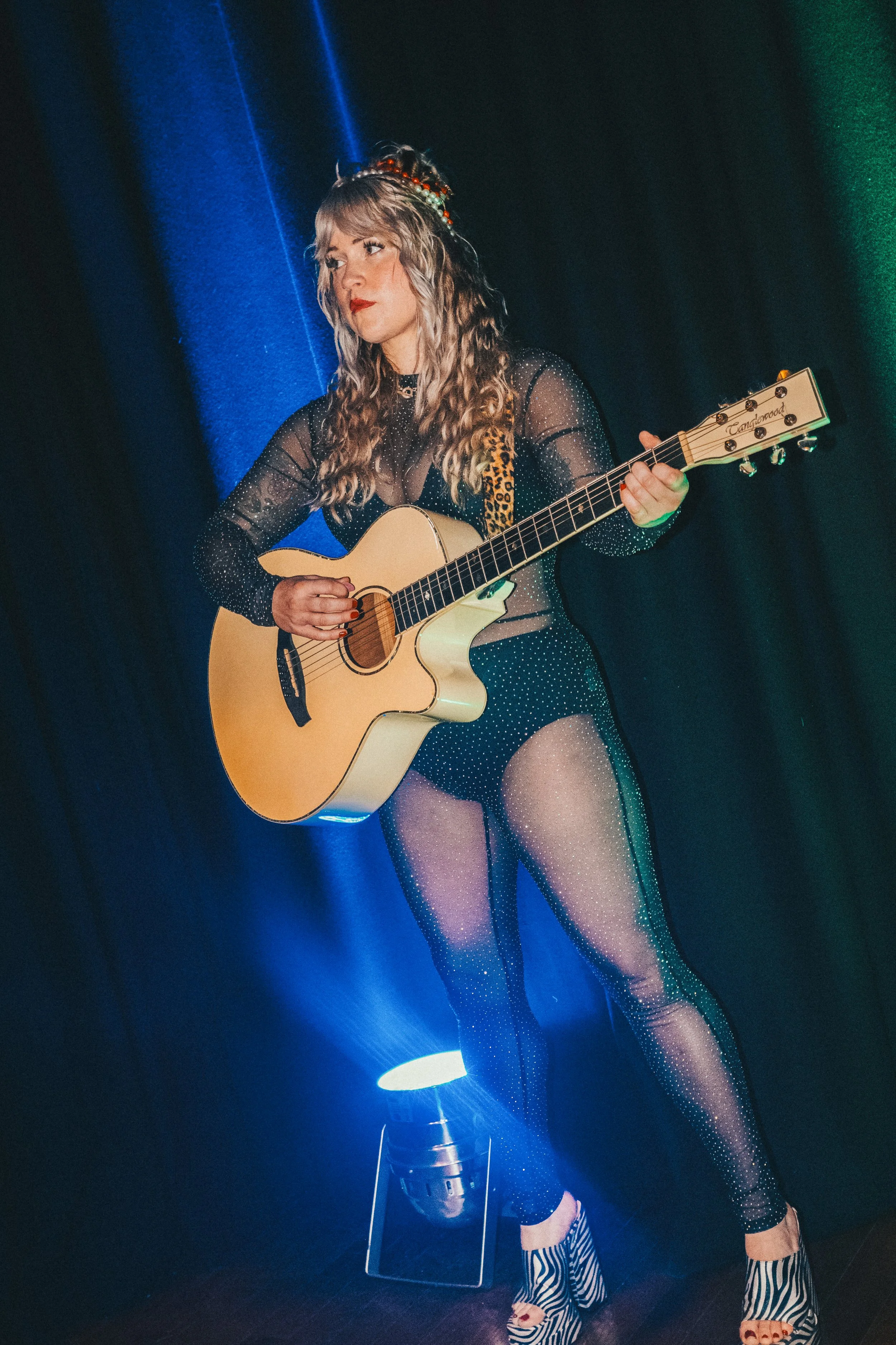 A woman with long curly blonde hair, wearing fishnet stockings, a black sparkly outfit, and zebra-patterned heels, is playing an acoustic guitar on a stage with colorful lighting.