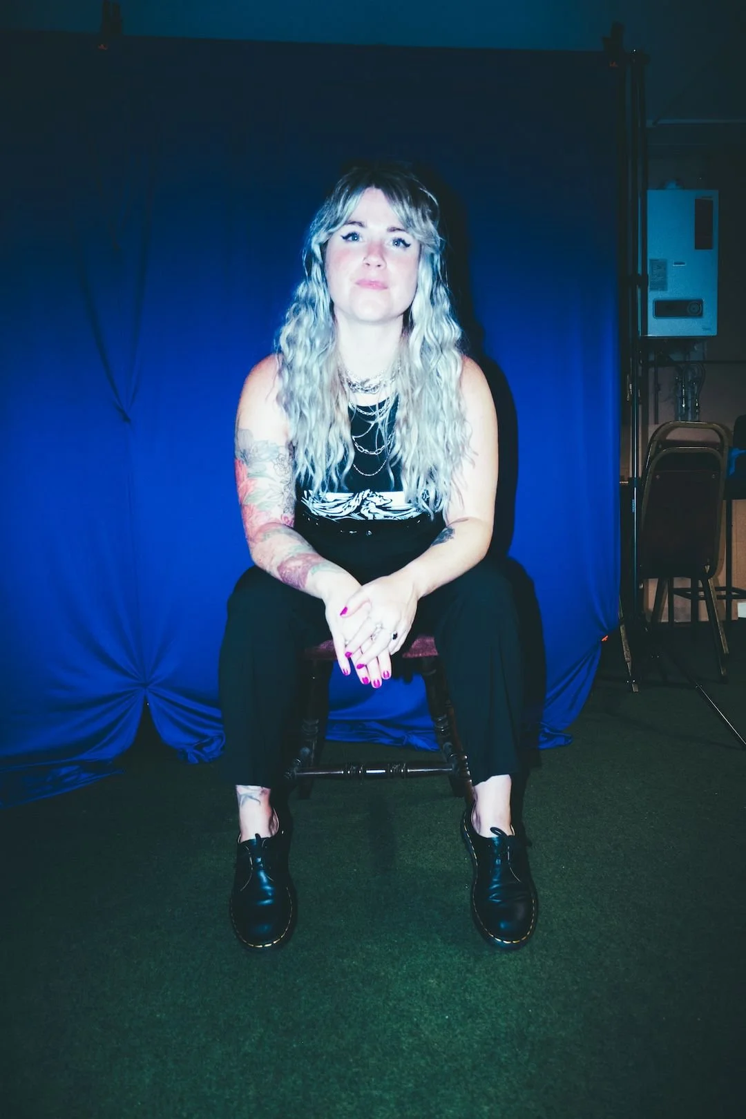 Woman with long wavy blonde hair, tattoos on her arms, wearing black outfit and black boots, sitting on a chair in front of a blue curtain, with a dark background and stage lighting.