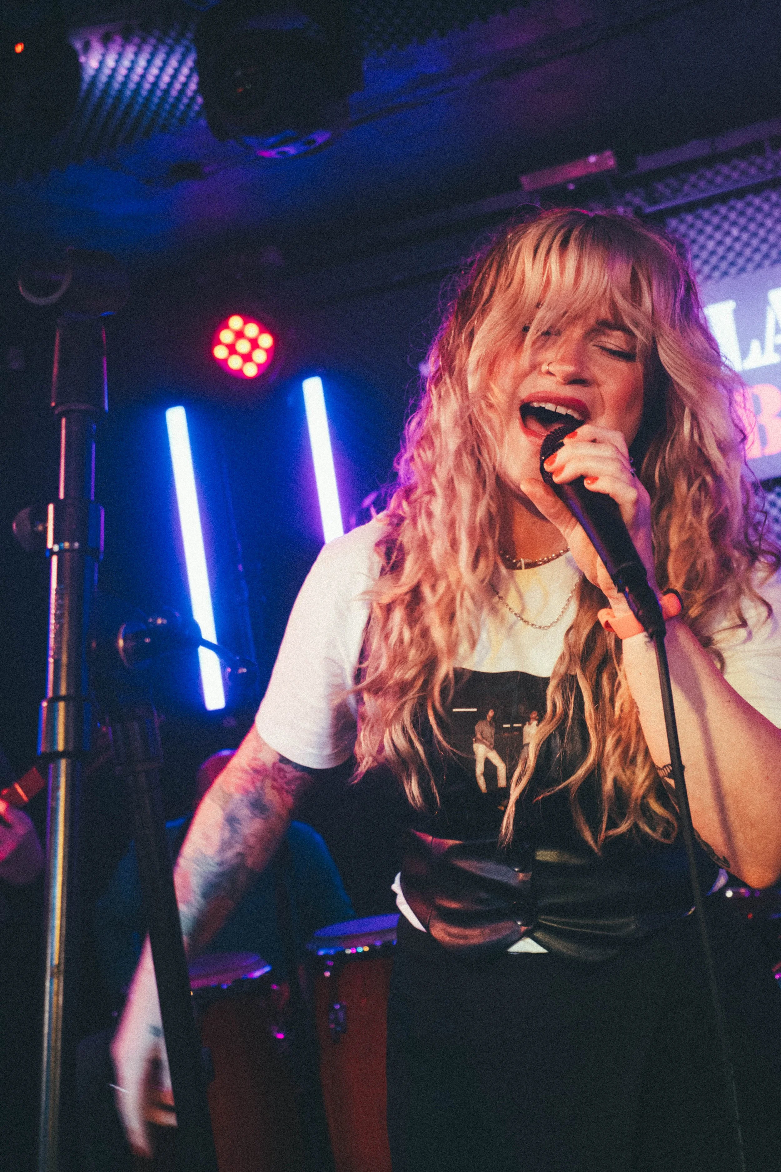 A woman with curly blonde hair sings passionately into a microphone on stage, illuminated by colorful neon lights.