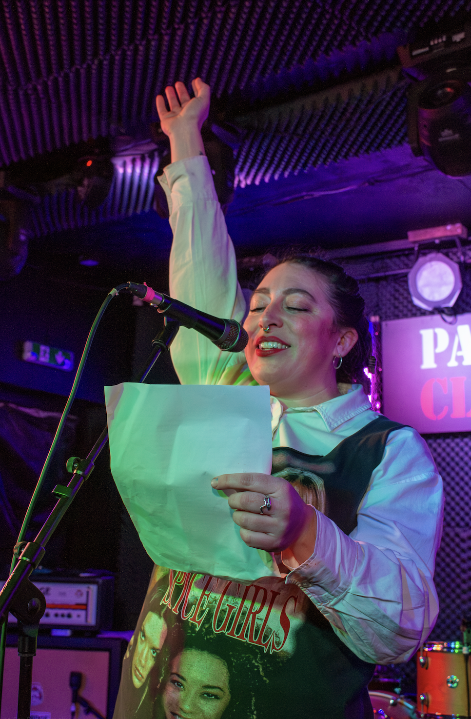 A woman singing or reading poetry into a microphone on stage, holding a sheet of paper, with her eyes closed and her right hand raised, in a colorful live music venue.