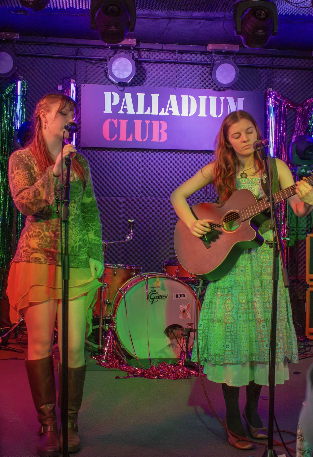 Two women performing music on stage at Palladium Club, one singing into a microphone and the other playing an acoustic guitar, with a drum set in the background and colorful lights.